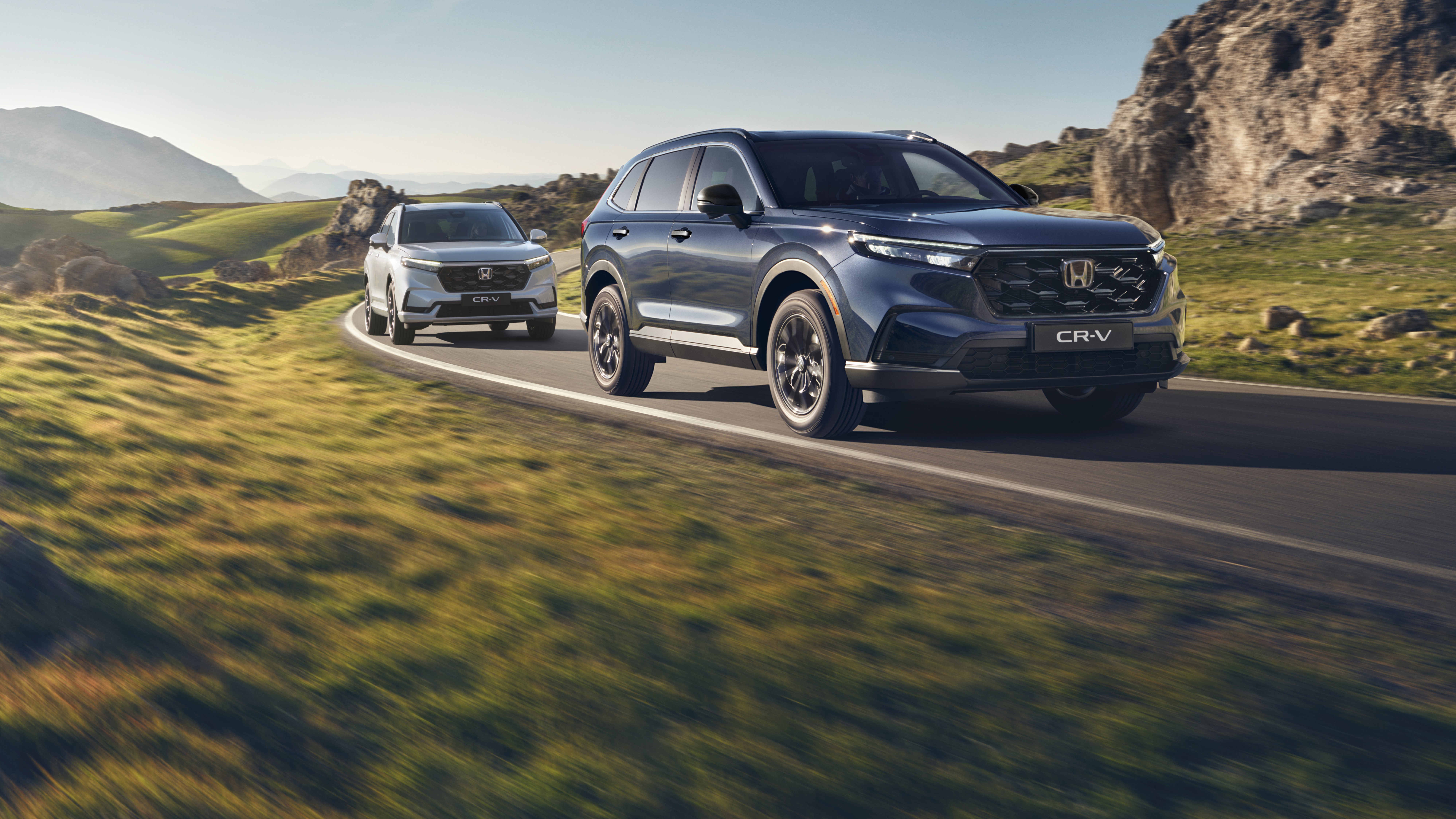 Un SUV Honda CR-V argenté roulant sur une route de montagne sinueuse avec un terrain rocheux et des montagnes en arrière-plan. Le véhicule est en mouvement, créant une sensation de vitesse et d'aventure dans ce paysage escarpé.