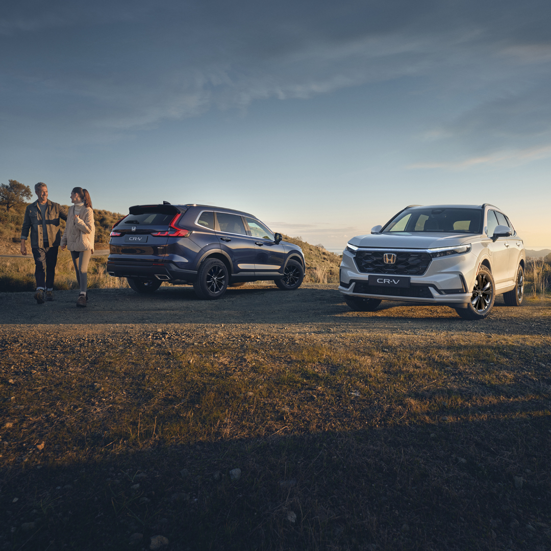 Deux SUV stationnés—un bleu foncé et un blanc—sont garés sur une clairière de terre dans un paysage rural à l'heure dorée, avec deux personnes marchant à proximité, des collines ondulantes et de hautes herbes en arrière-plan sous un ciel dégagé.