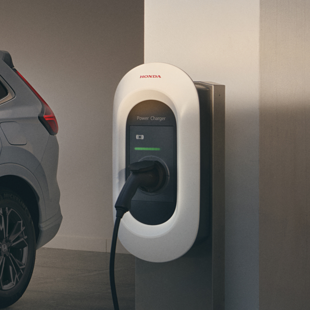 Une borne de recharge pour véhicules électriques Honda fixée au mur, avec un câble de recharge blanc branché à un CR-V Plug-in hybride blanc, vue de profil