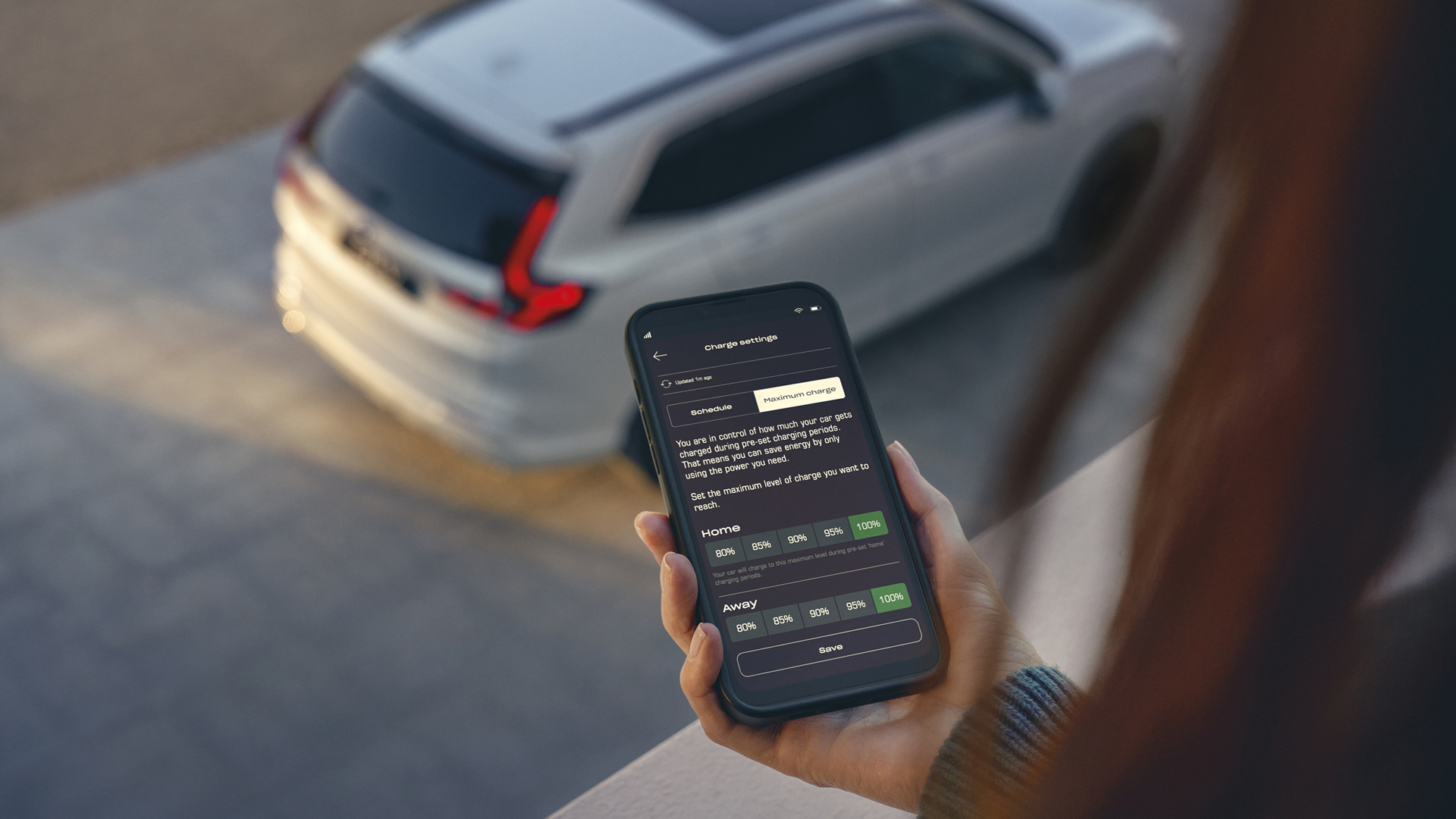 Une personne tient un smartphone affichant une application de gestion de la recharge du véhicule, tandis qu'un SUV blanc est garé en arrière-plan.