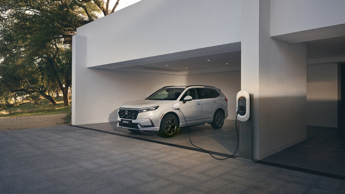 Un CR-V plug-in hybride blanc est garé à l'entrée d'une maison et est en train de charger à une borne de recharge.