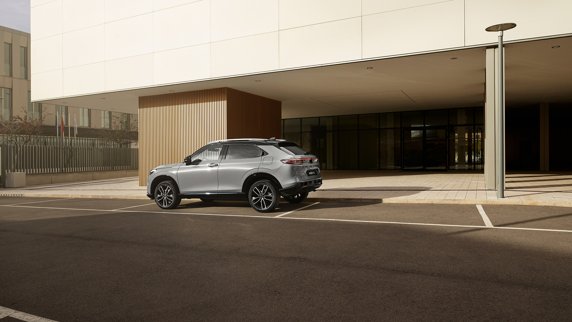 Un Honda HR-V argenté est stationné sur une place extérieure devant un bâtiment moderne aux parois vitrées et aux panneaux verticaux en bois, photographié de jour en vue arrière trois quarts.