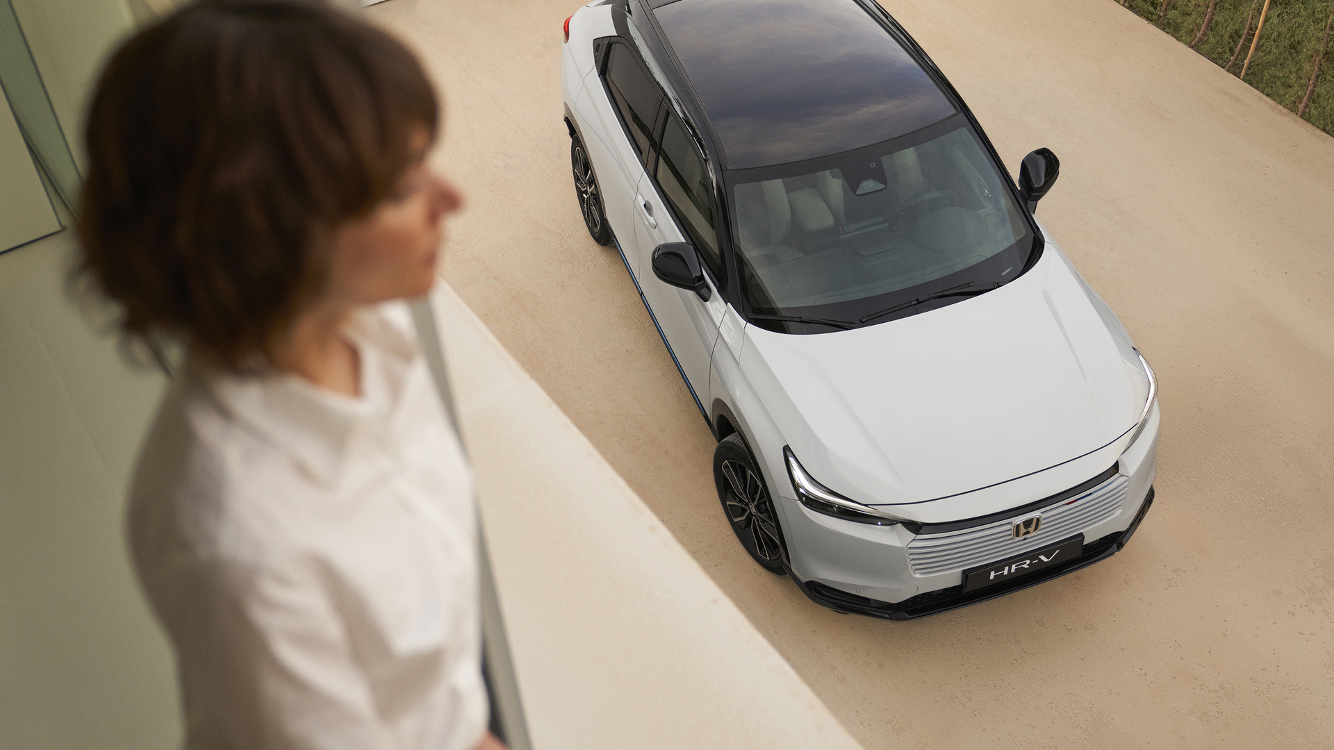 Vue de dessus d’un Honda HR-V blanc à toit noir, stationné sur une allée claire, tandis qu’une personne floue se tient à l’intérieur au premier plan et regarde vers le véhicule.