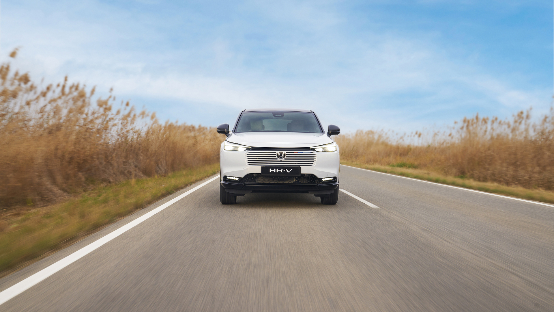Un Honda HR-V blanc roule vers l’objectif sur une route à deux voies, au milieu de hautes herbes dorées sous un ciel bleu.