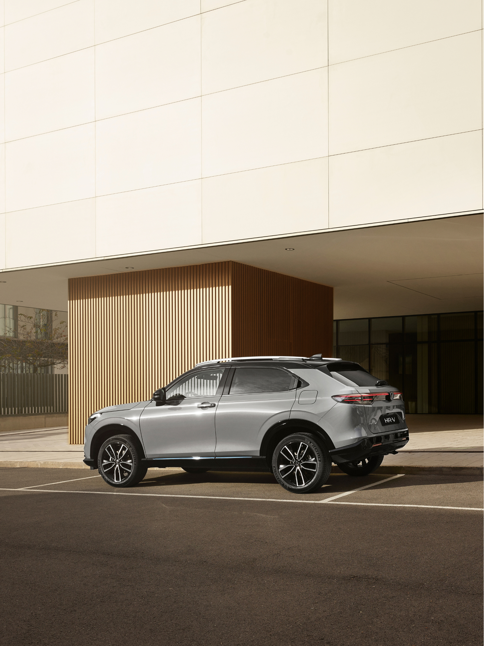 Un Honda HR-V argenté est stationné sur une place extérieure devant un bâtiment moderne aux parois vitrées et aux panneaux verticaux en bois, photographié de jour en vue arrière trois quarts.