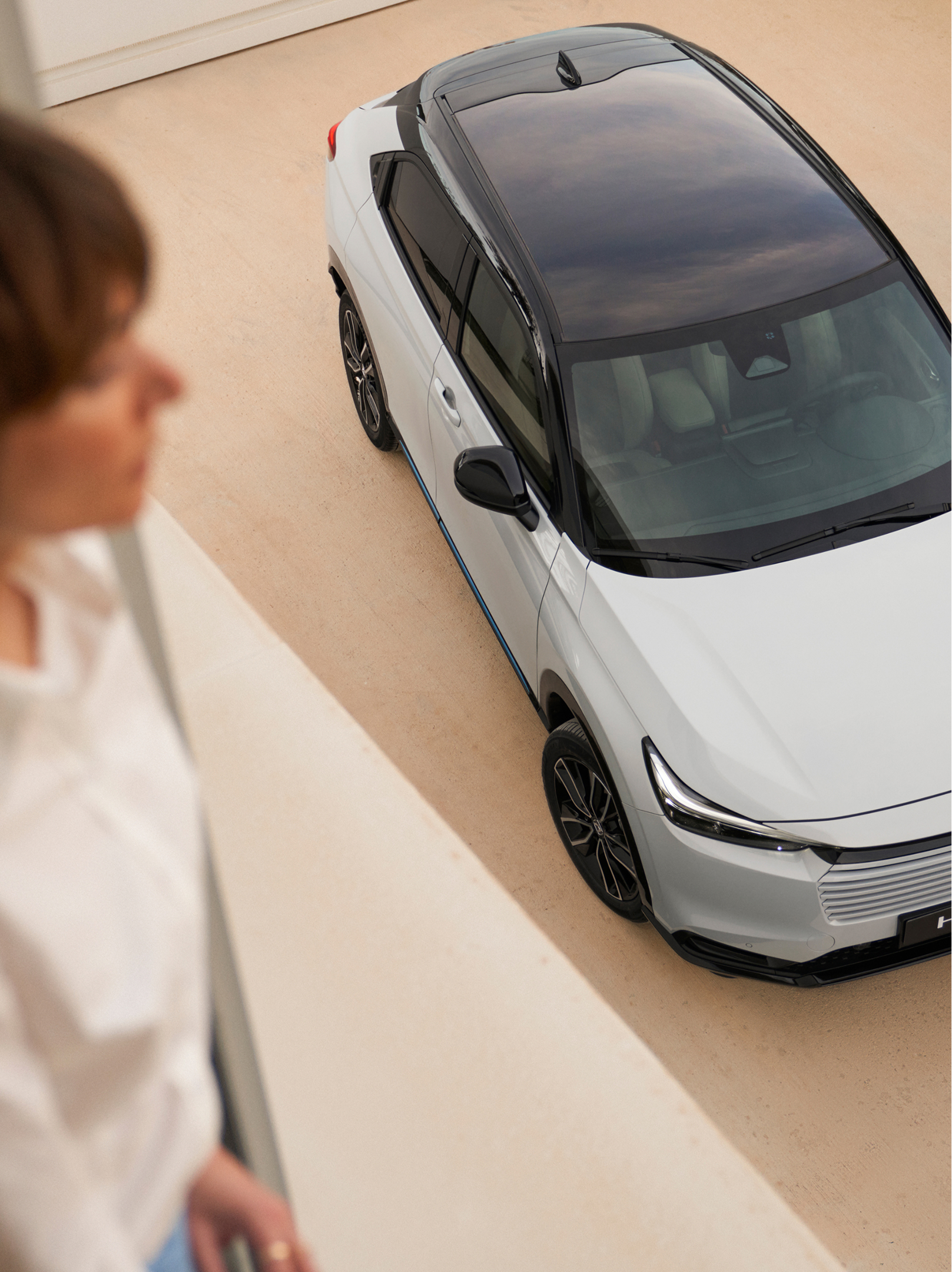 Vue de dessus d’un Honda HR-V blanc à toit noir, stationné sur une allée claire, tandis qu’une personne floue se tient à l’intérieur au premier plan et regarde vers le véhicule.