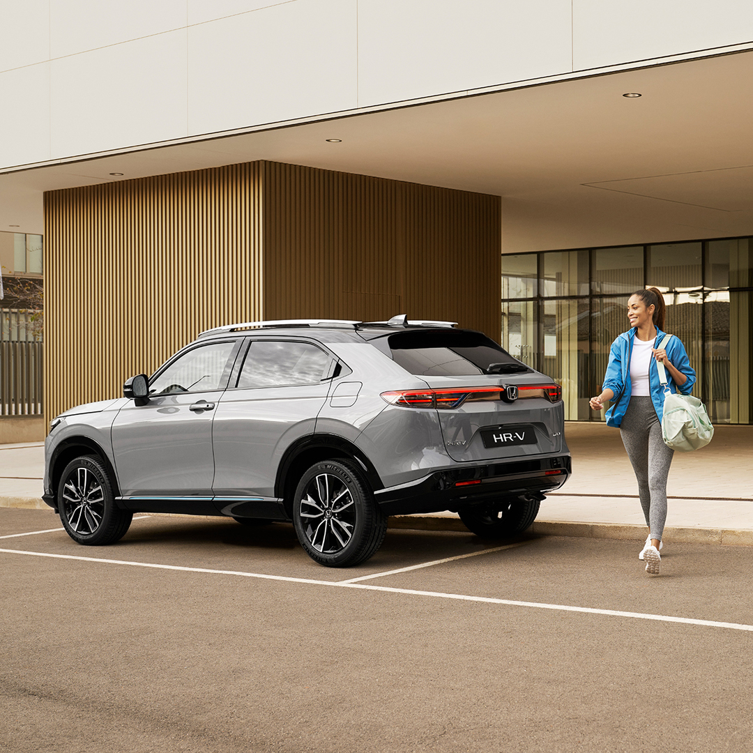 Un Honda HR-V argenté est stationné devant un bâtiment moderne, tandis qu’une femme passe avec un sac, évoquant un usage urbain pratique au quotidien.