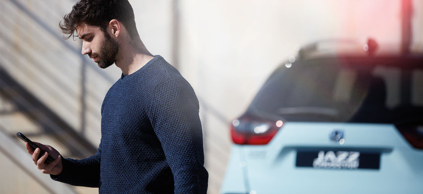 Homme regardant l'application My Honda Plus avec la Honda Jazz Hybrid Crosstar derrière. 