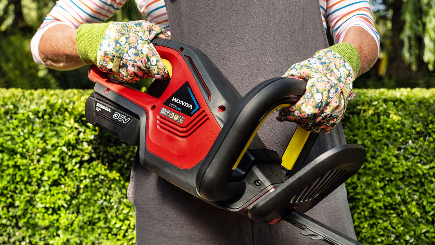 Modèle tenant un taille-haies à batterie Honda par la poignée dans un jardin.