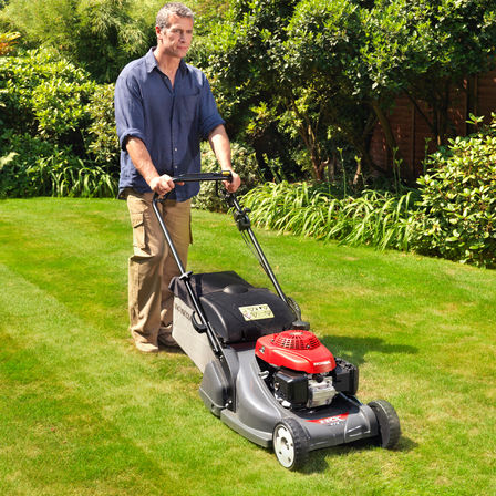 Tondeuse à gazon, de trois quarts face, tournée vers la droite, utilisée par un démonstrateur, dans un jardin.