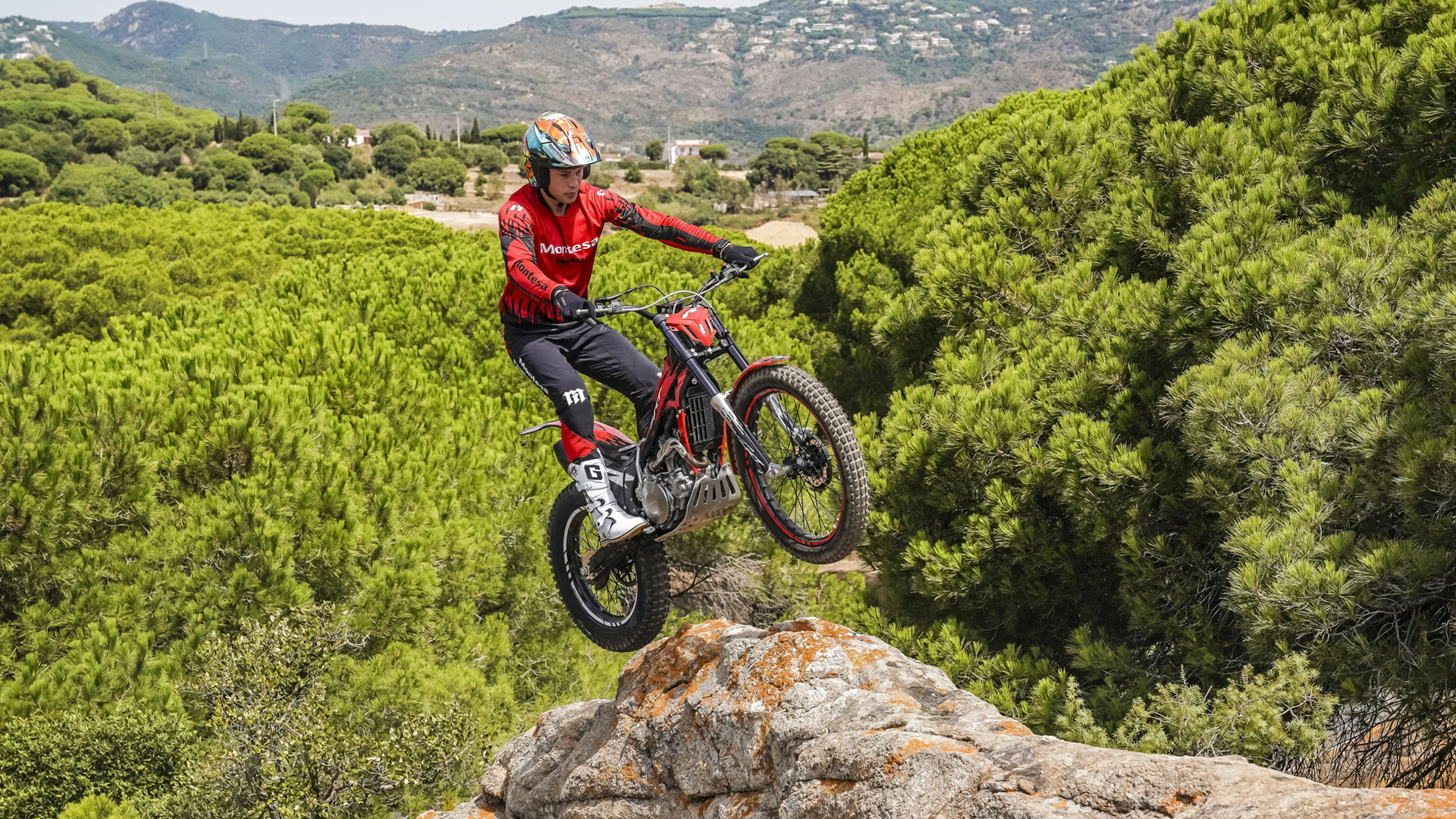 Pilote sautant d’une corniche rocheuse avec une Montesa Cota 4RT 260R rouge, au-dessus d’un paysage de pins.