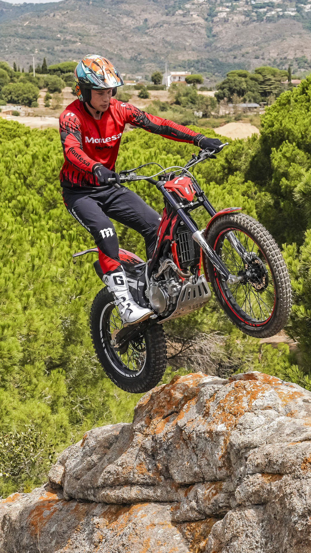 Pilote sautant d’une corniche rocheuse avec une Montesa Cota 4RT 260R rouge, au-dessus d’un paysage de pins.