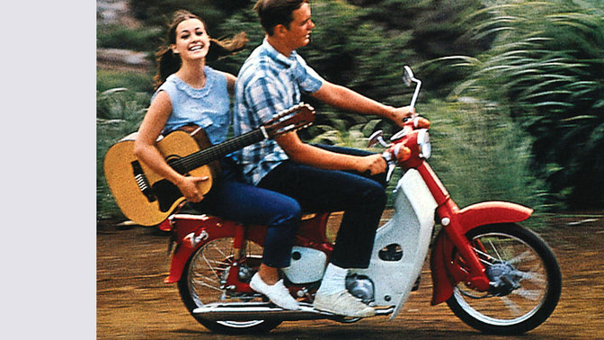 Honda Cub avec conducteur et passager jouant de la guitare. Chemin en terre. Vue latérale côté droit.
